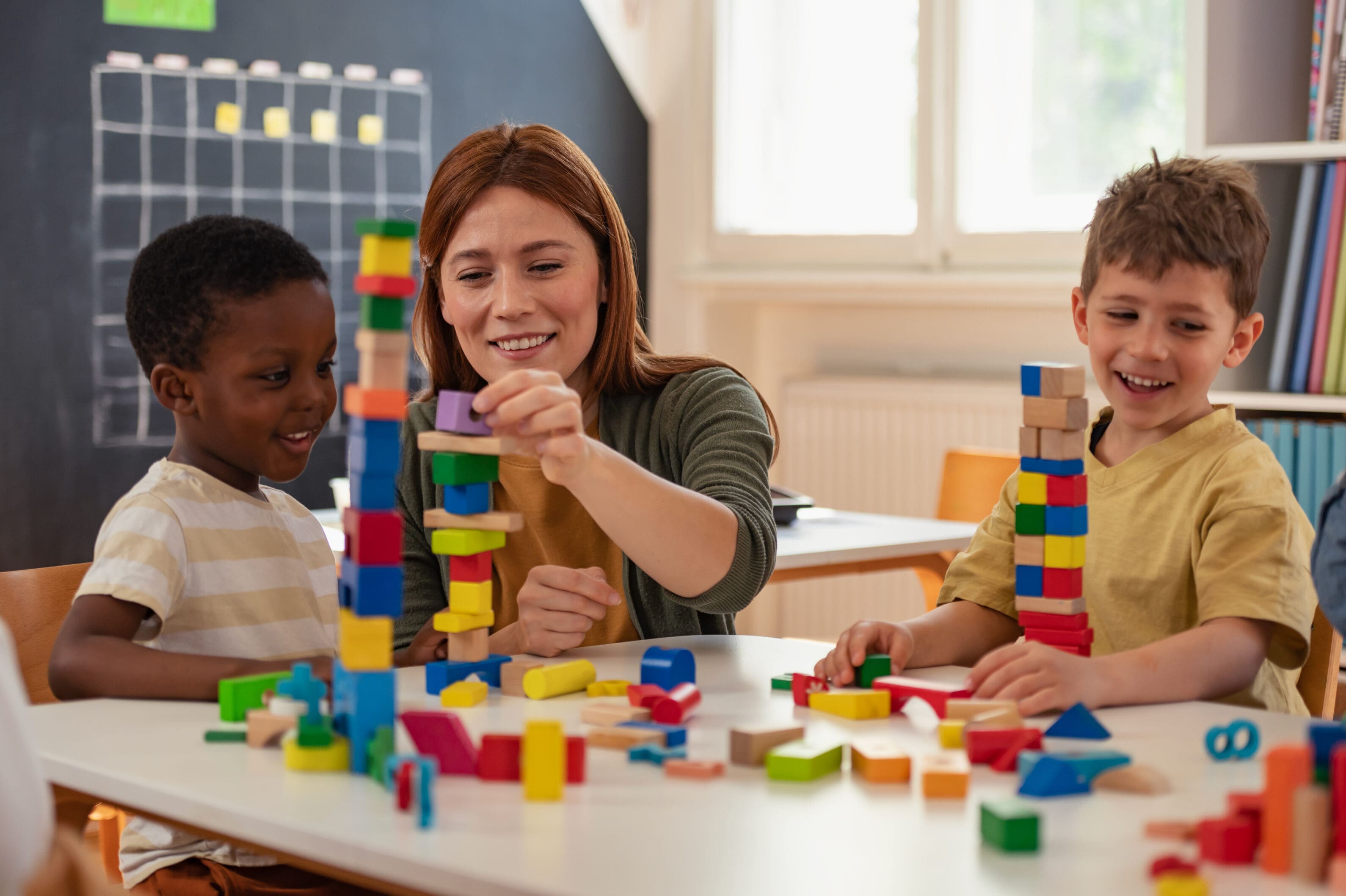 Diverse preschool children play with colorful building blocks at a classroom table, guided by a smiling teacher in a creative, inclusive early learning environment.